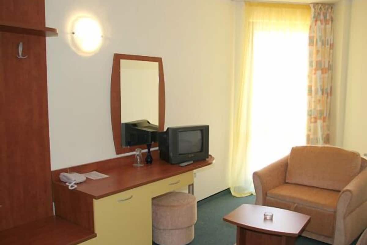 A compact hotel room with a wooden desk, a television, a small table, a chair, and a window with curtains.