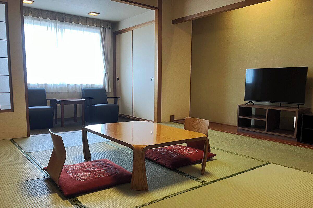 Une pièce de style japonais traditionnel, avec un sol recouvert de tatamis, une basse table en bois entourée de coussins, un téléviseur et une porte coulissante donnant sur un espace salon.