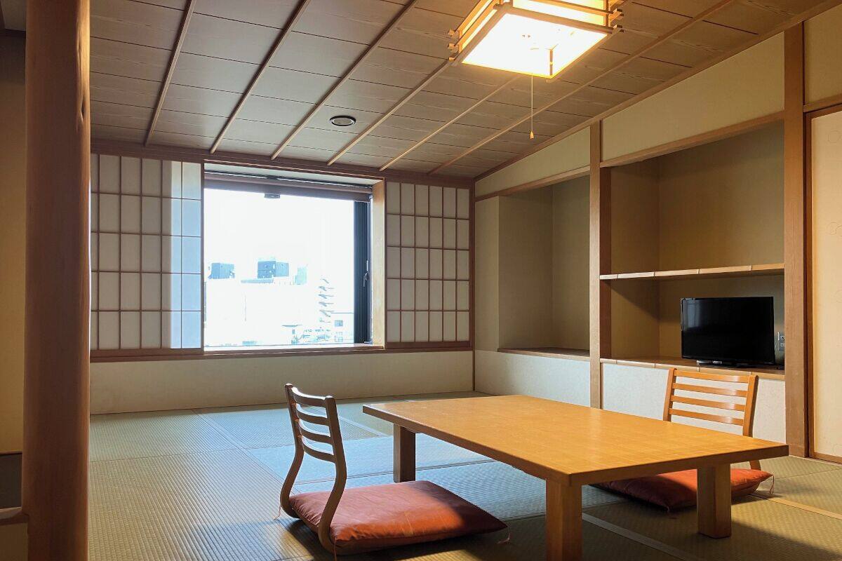 Une pièce de style japonais traditionnel, avec un sol recouvert de tatamis, une table basse, deux chaises en bois, un téléviseur posé sur une étagère et une porte coulissante donnant sur une fenêtre avec vue sur les bâtiments.