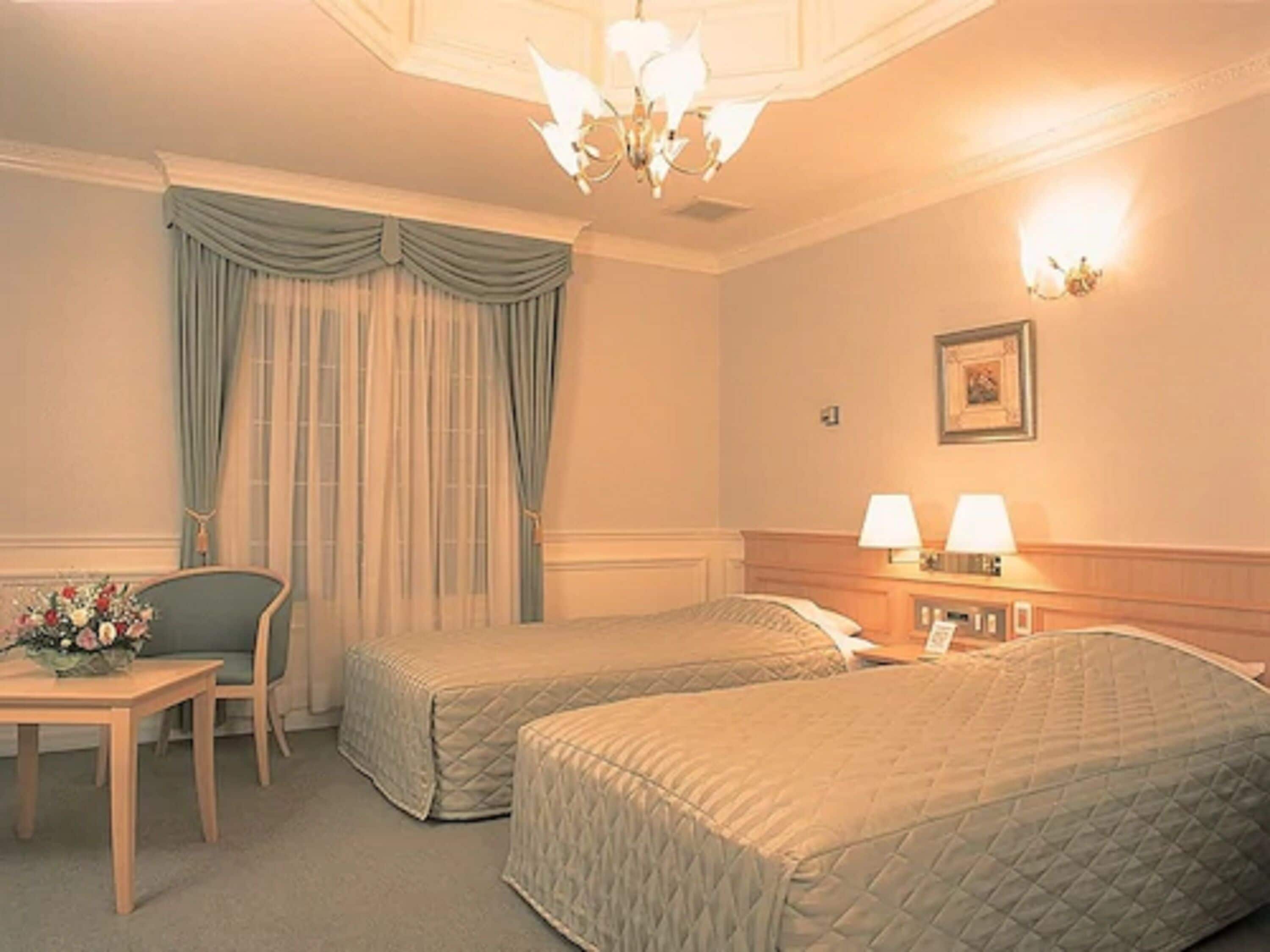 Une chambre d’hôtel avec deux lits simples, un bureau avec une chaise, une table agrémentée d’un arrangement floral et un lustre.