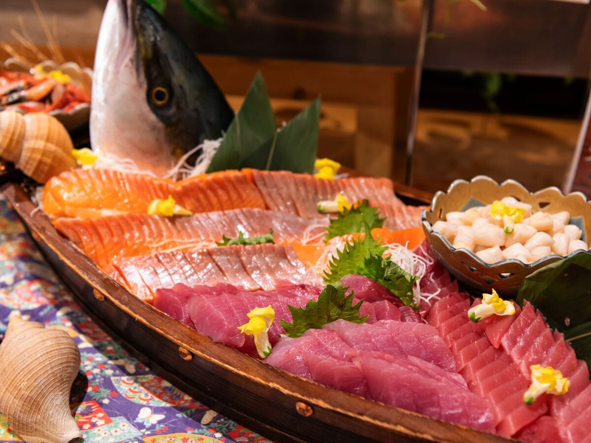 A wooden tray with various types of raw fish, including salmon and tuna, garnished with green leaves and yellow flowers.