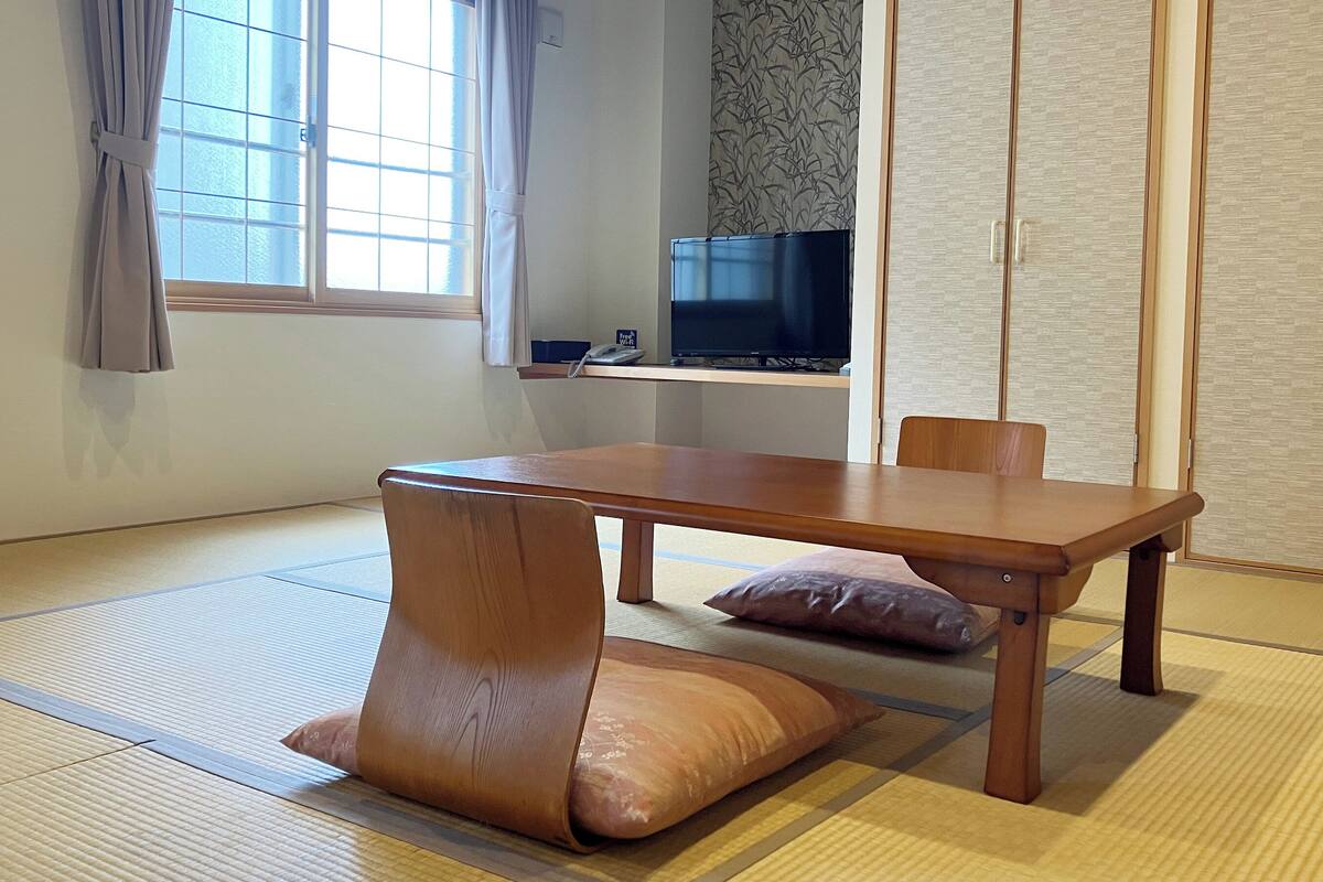 Une pièce de style japonais traditionnel, avec un sol recouvert de tatamis, une table basse, des coussins, un téléviseur et des portes coulissantes.