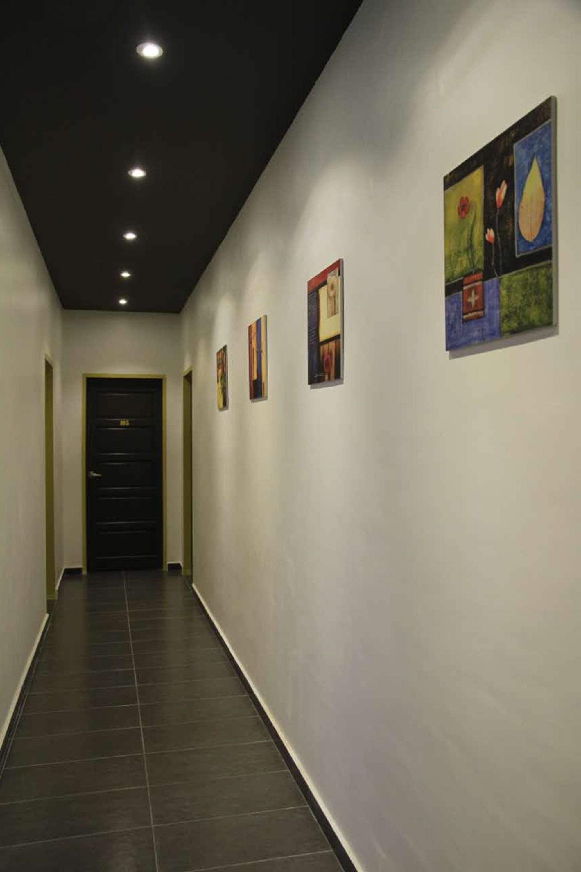 A hallway with a black door at the end, recessed lighting, and framed artwork on the walls.