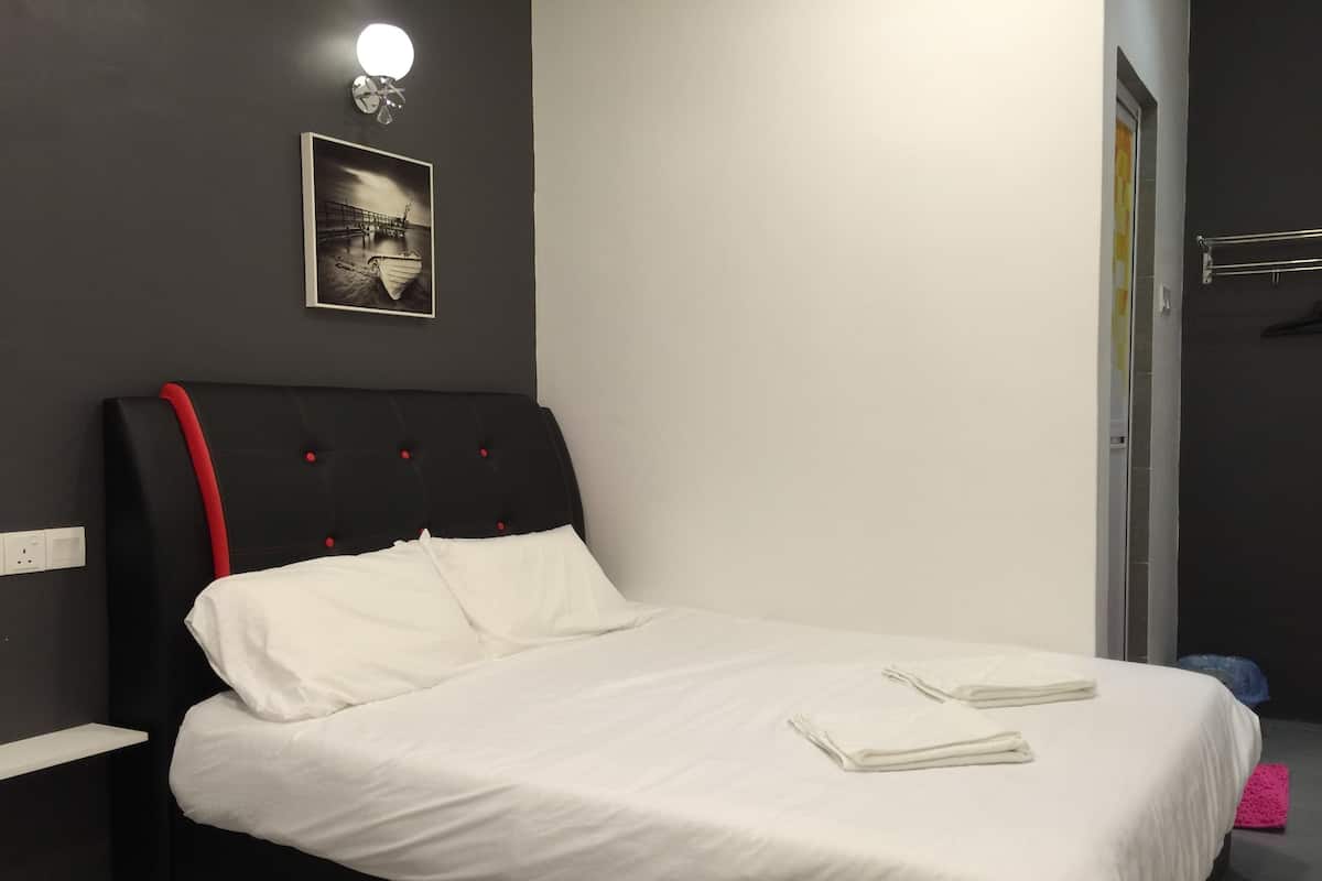 Una cama bien hecha con sábanas blancas y dos toallas dobladas, cabecera negra con detalles rojos, lámpara de pared y un cuadro enmarcado sobre la cama.