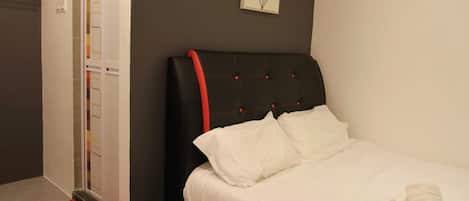 Un lit bien fait, avec du linge de lit blanc et un élément décoratif rouge, un tapis rouge au pied du lit et un tableau encadré au mur.