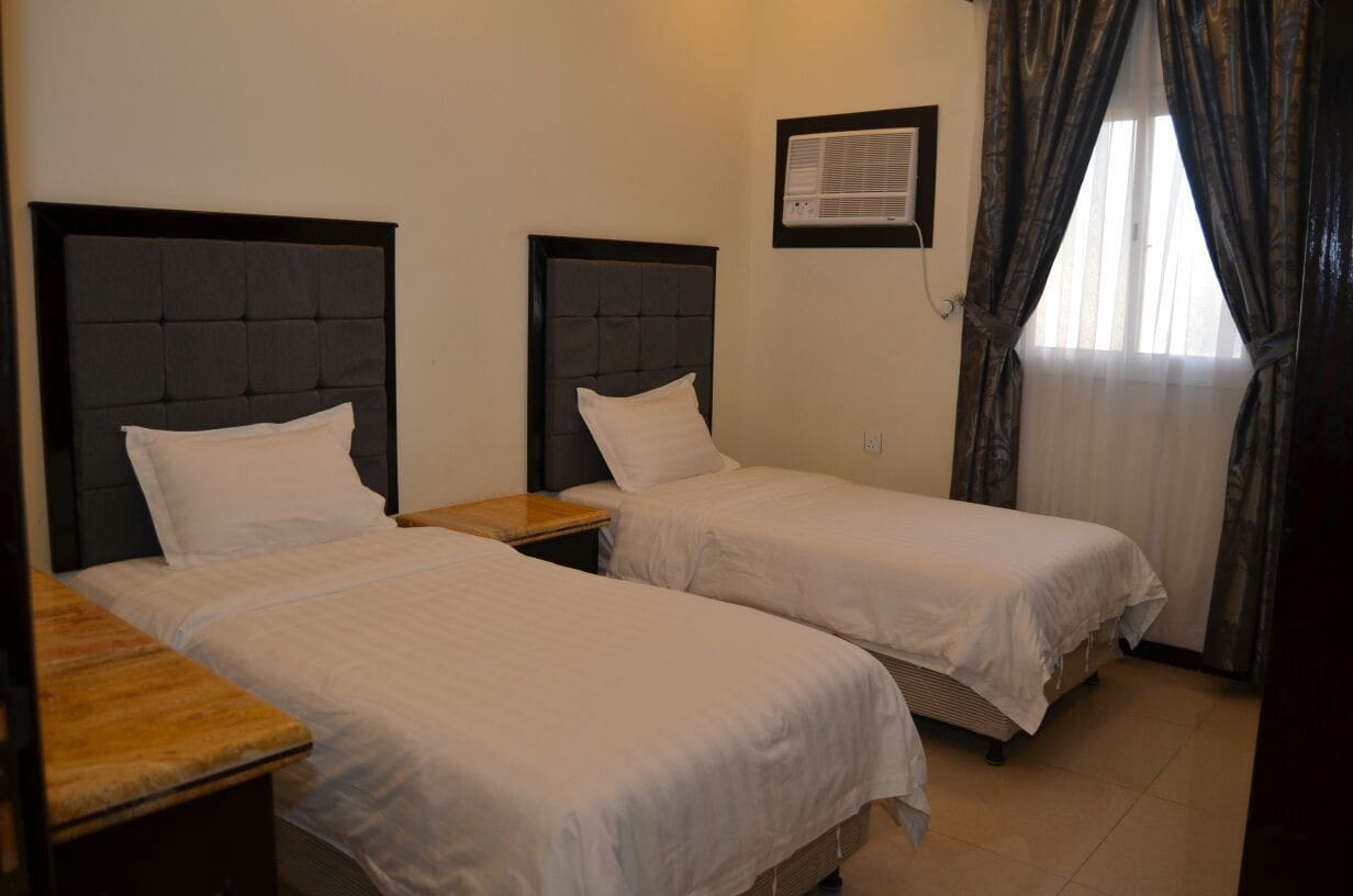 A room with two single beds, a wooden nightstand, a wall-mounted air conditioner, and a window with curtains.