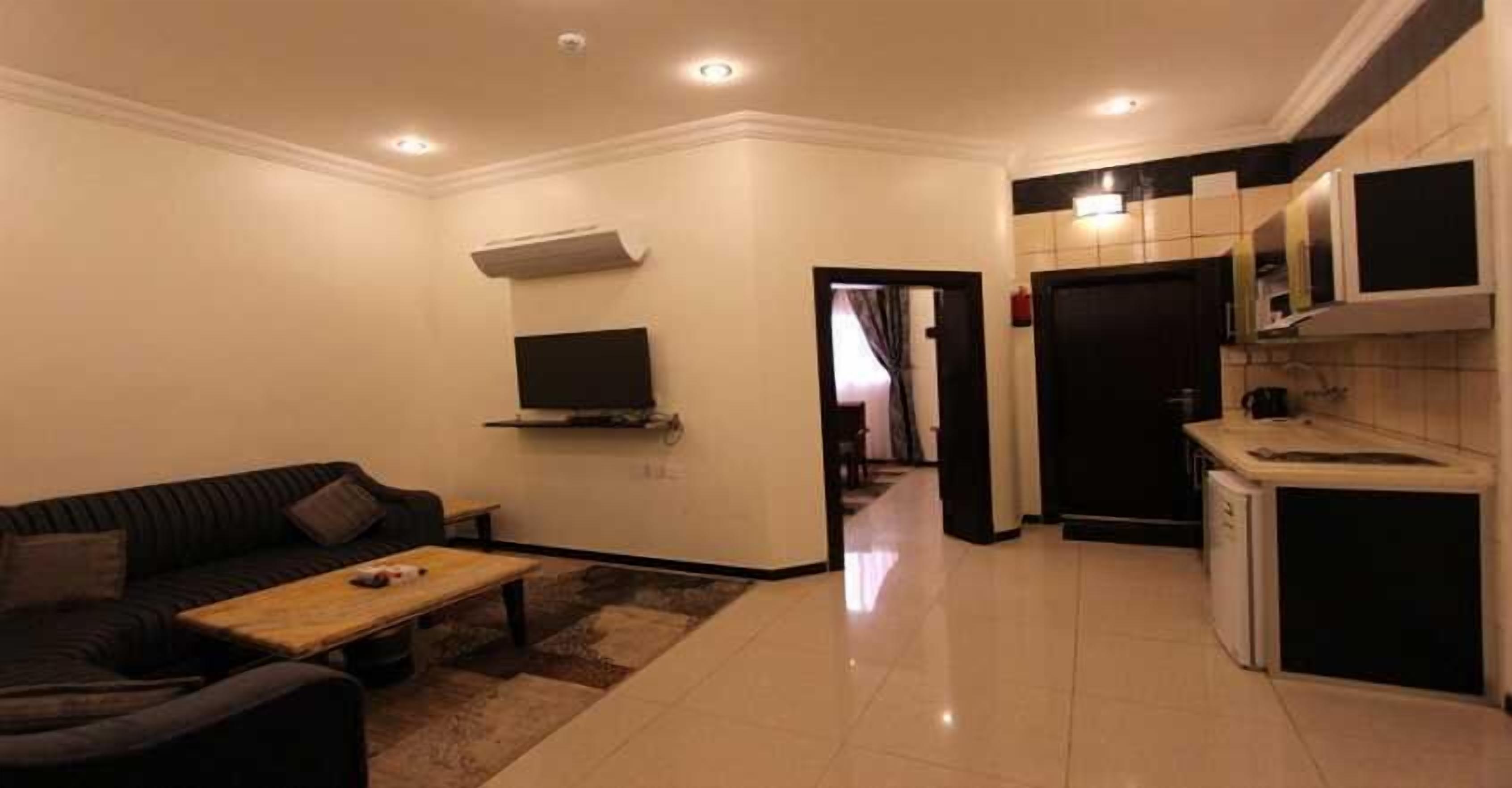 Una sala de estar moderna con un sofá, una mesa de centro y un televisor. Hay una zona de cocina con microondas y refrigerador.