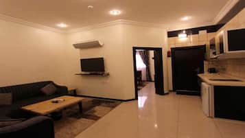 Una sala de estar moderna con un sofá, una mesa de centro y un televisor. Hay una zona de cocina con microondas y refrigerador.