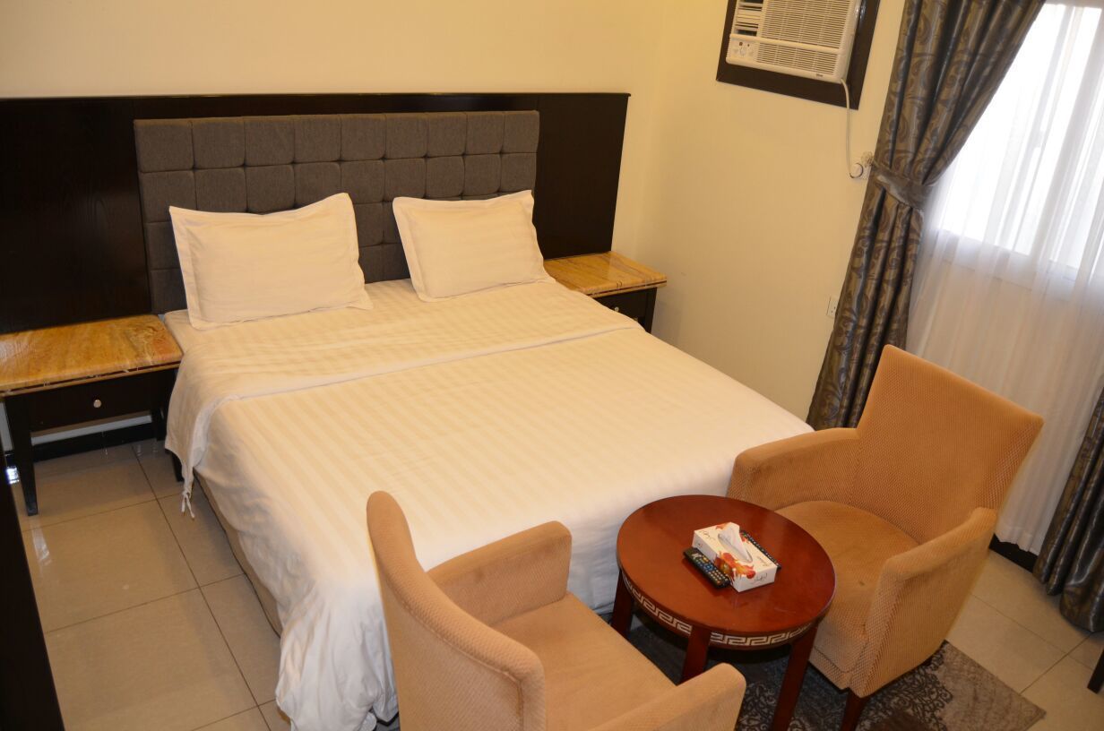 A hotel room with a large bed, two pillows, a wooden nightstand, a small round table with a remote control, and two armchairs.