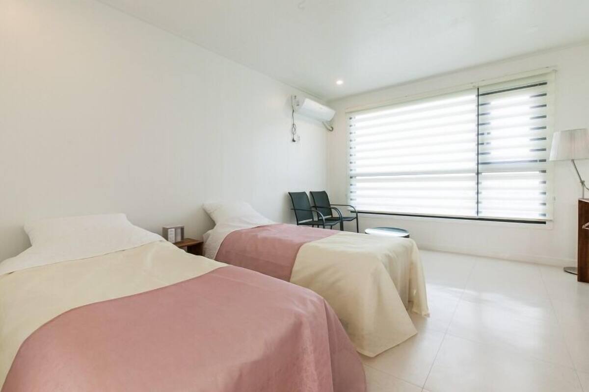 Habitación de hotel con dos camas, una mesita, una silla y una ventana con persianas.