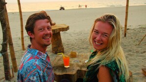 Ein Holztisch mit einem Getränk, auf einem Sandstrand mit einer Holzkonstruktion im Hintergrund.
