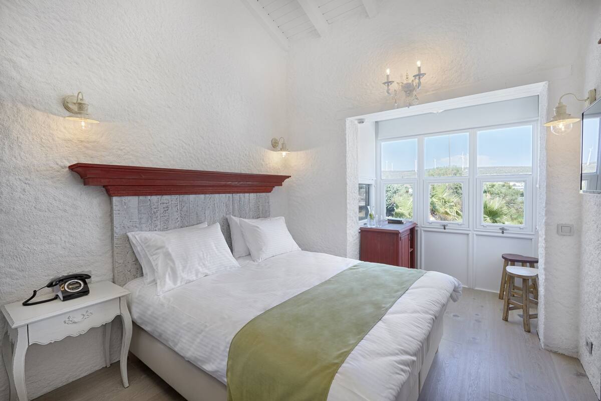 A bedroom with a bed, bedside table, nightstand, and a window with a view.