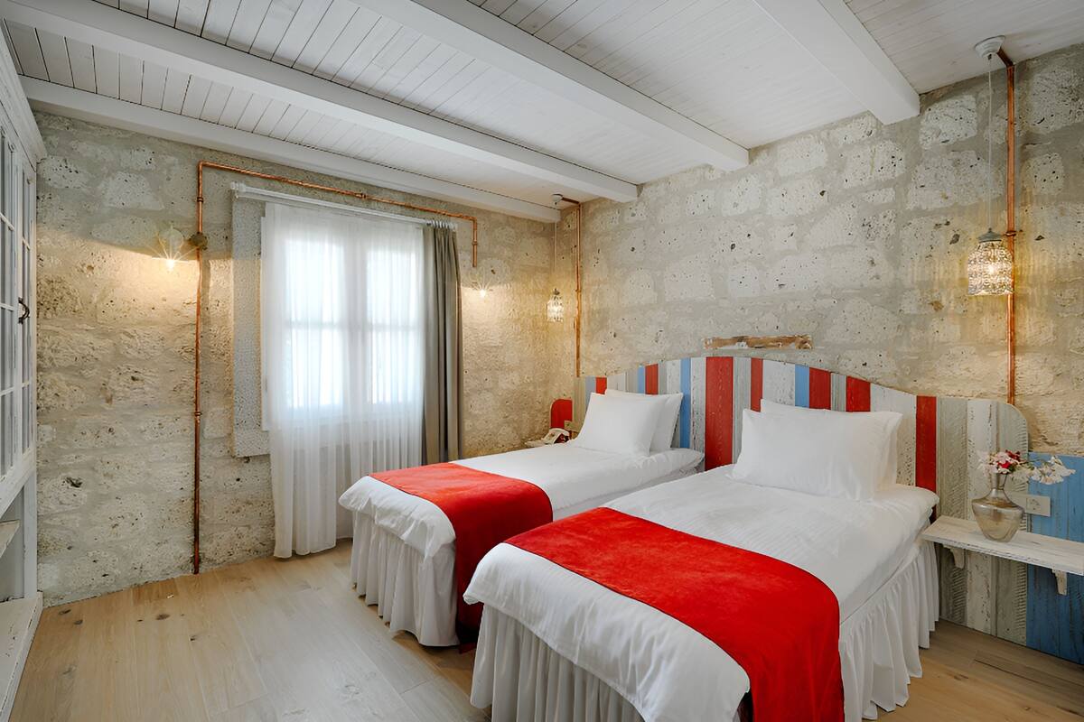 A room with two beds, a wooden floor, a stone wall, a window with curtains, and a hanging light fixture.