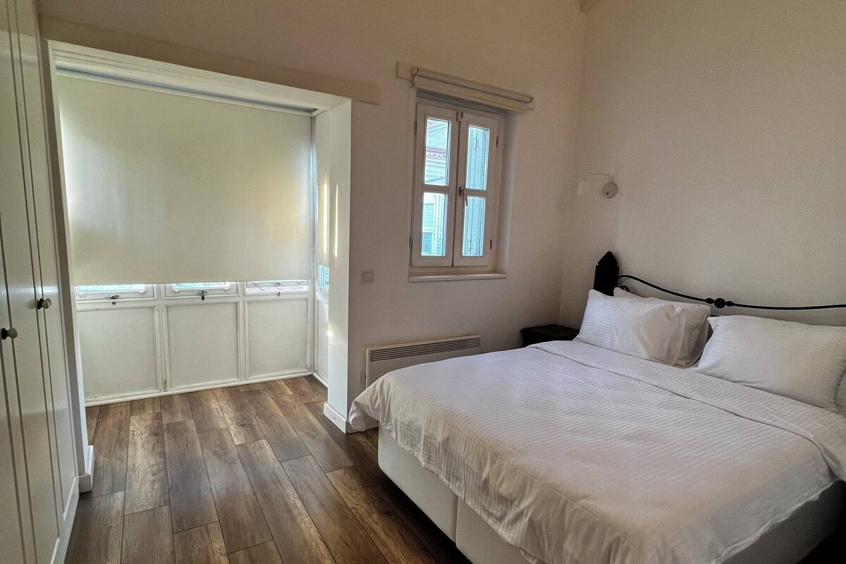 A bedroom with a wooden floor, a bed with white bedding, a window with blinds, and a ceiling fan.