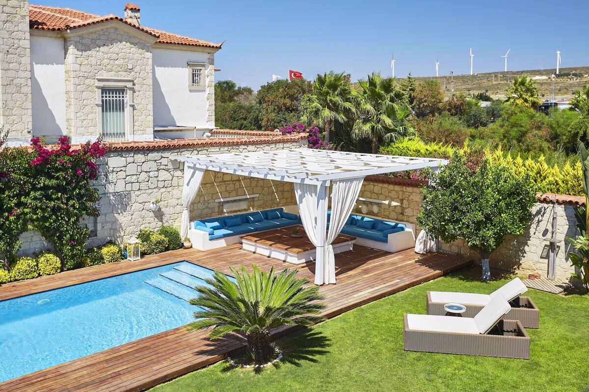 A pool area with a wooden deck, lounge chairs, and a pergola with seating.