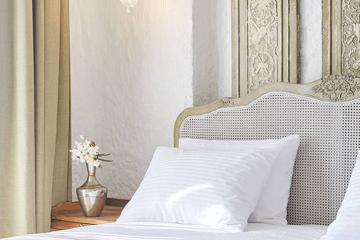 A bedroom with a bed, white bedding, a wall-mounted lamp, a nightstand with a vase of flowers, and ornate doors.