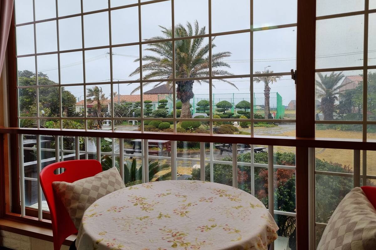 Ein Zimmer mit einem großen Fenster, einem runden Tisch mit einer blumigen Tischdecke und roten Stühlen.