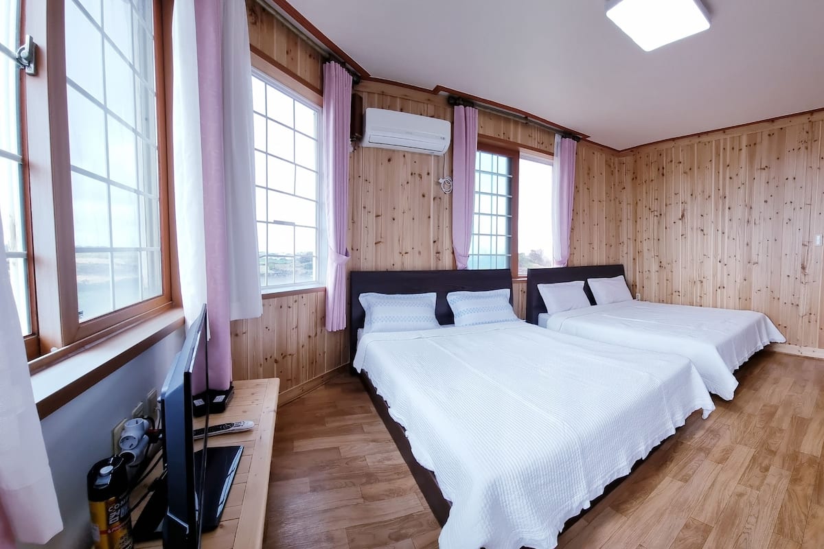 Ein Zimmer mit zwei Betten, Holzwänden, einem Fenster mit Vorhängen, einem Schreibtisch mit Stuhl und einer Leiter.