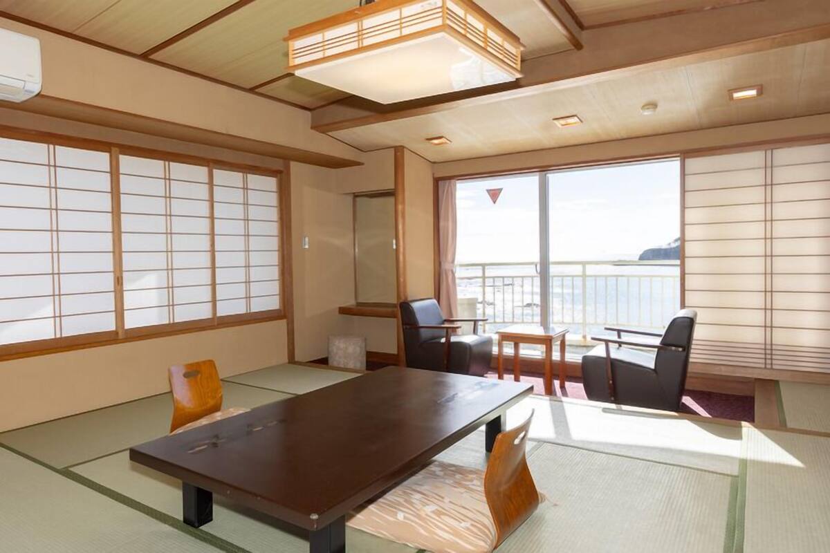 A traditional Japanese room with tatami flooring, a low table, chairs, a sliding door, and a view of the sea.
