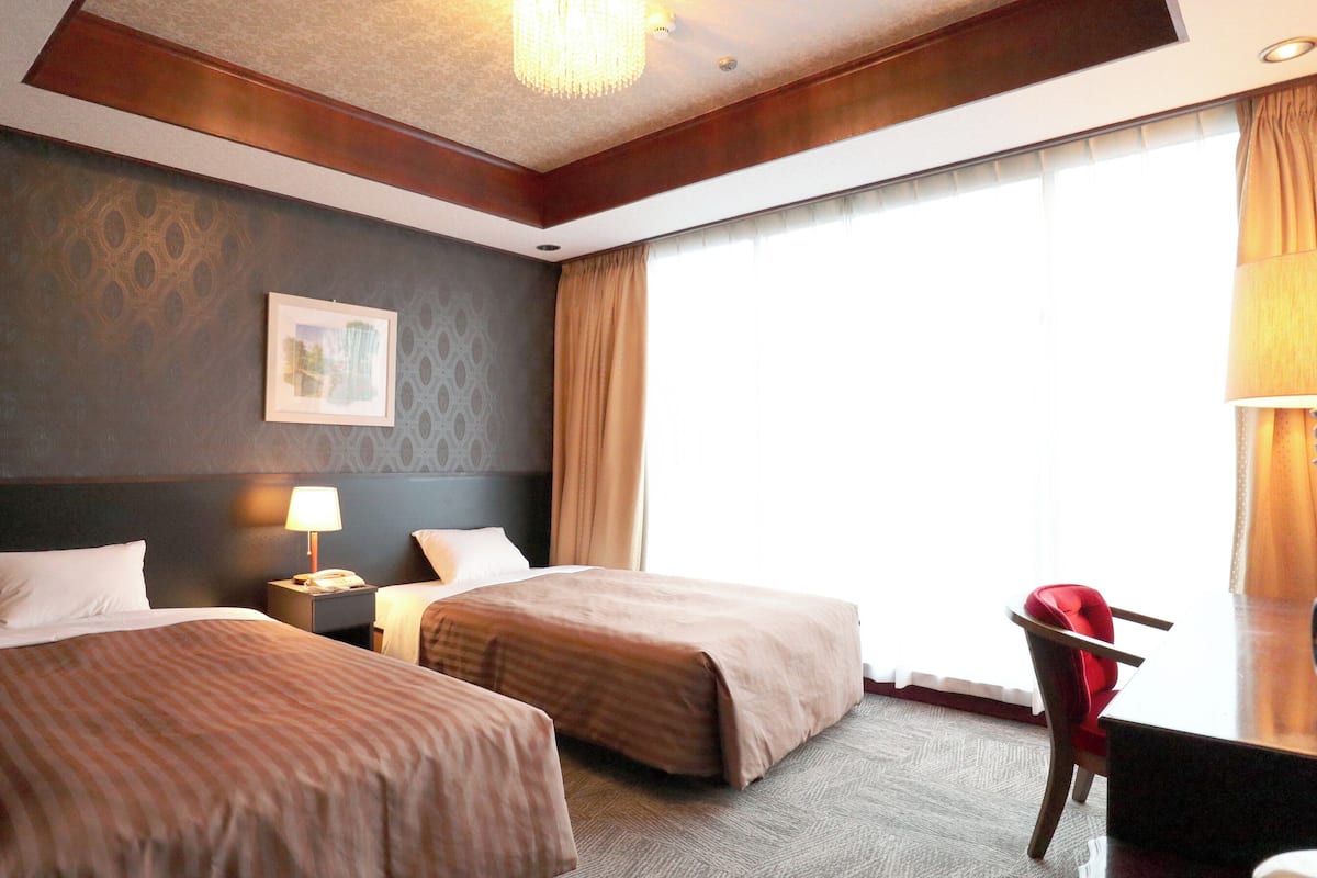 Habitación de hotel con dos camas, un escritorio, una silla, una lámpara y un ventanal grande.