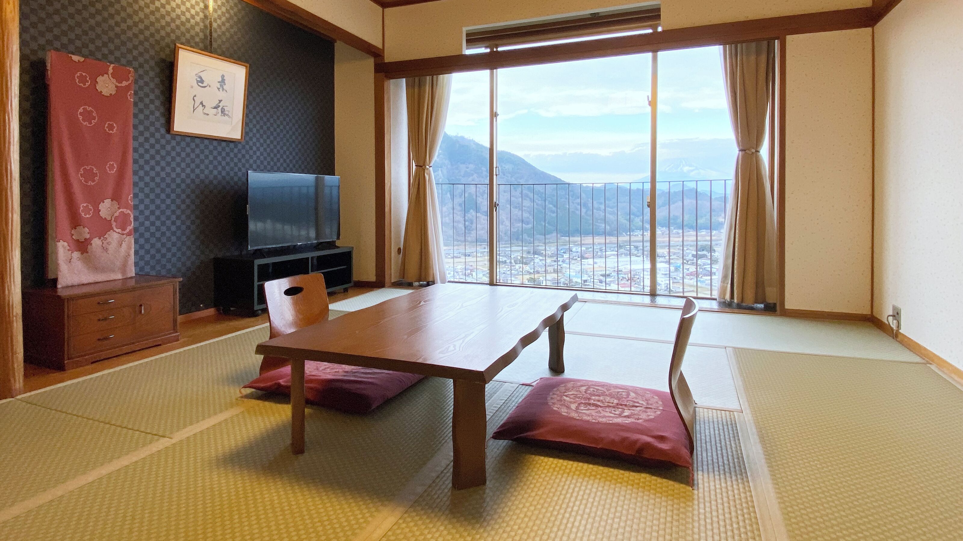A traditional Japanese room with tatami flooring, a low table, cushions, a TV, and a view of a mountain.