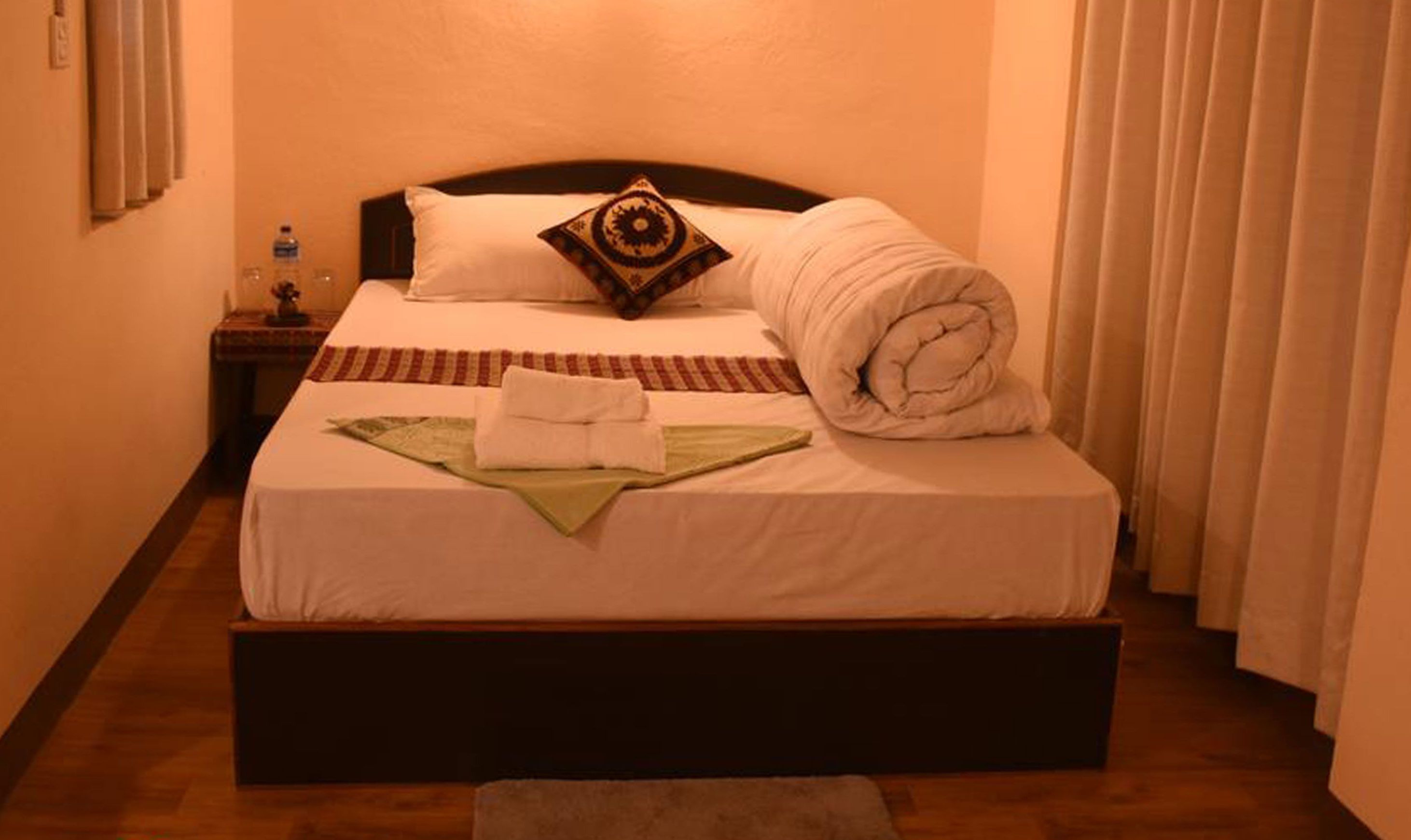 A neatly made bed with a pillow, a folded blanket, and a rolled-up blanket on a wooden platform bed in a hotel room.