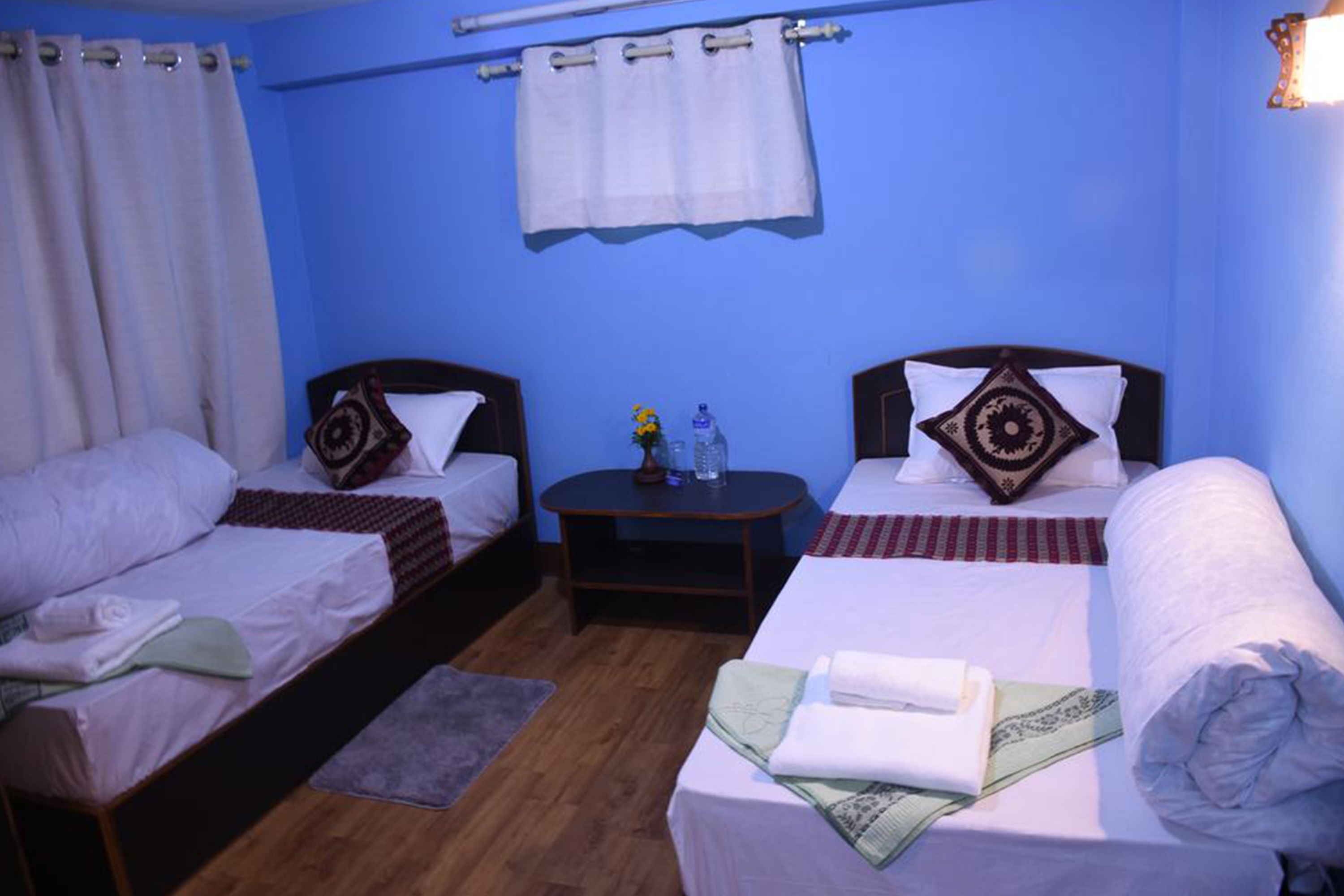 A room with two single beds, a round table with a bottle and glasses, and a large bed with a rolled-up blanket.