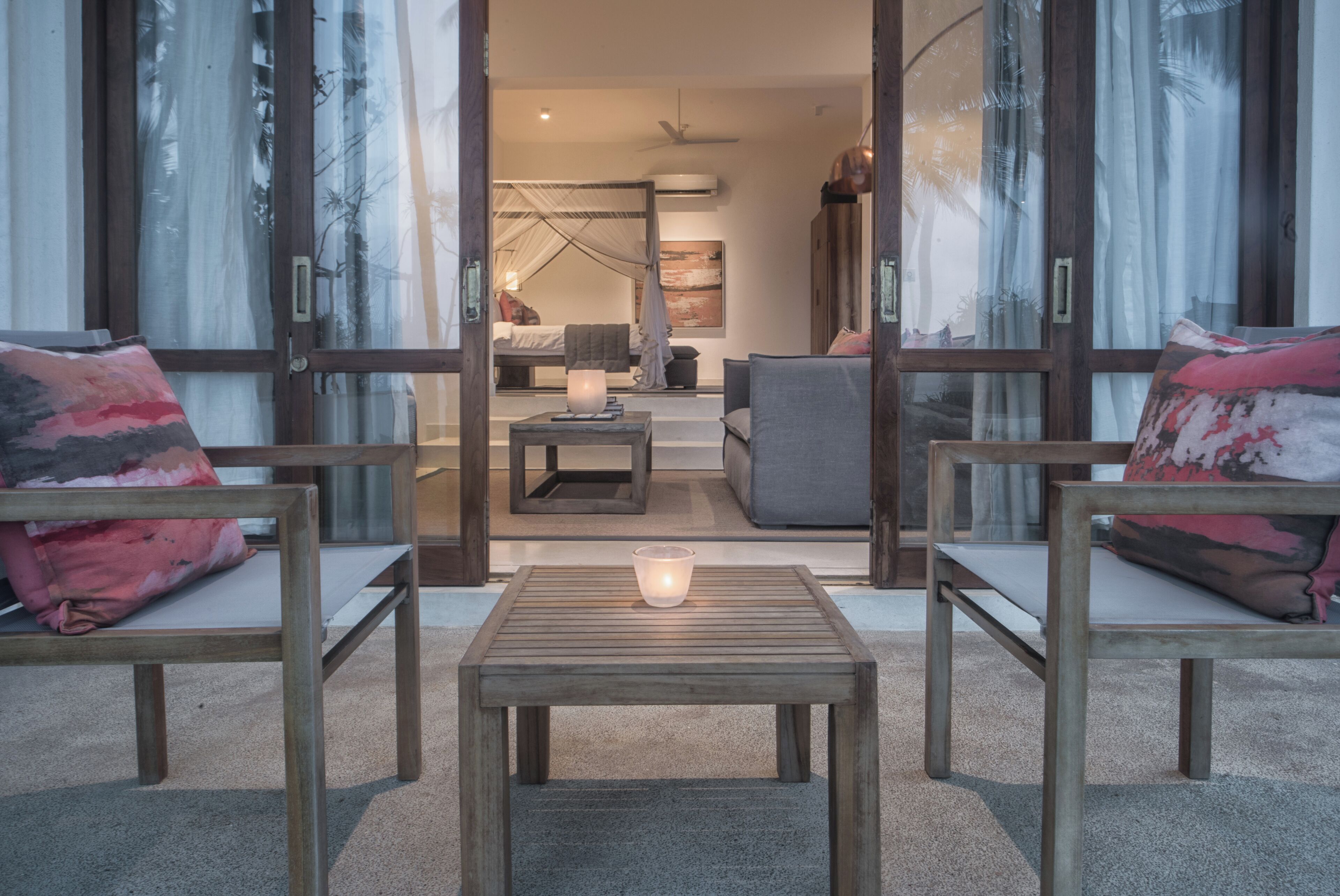 Ein modernes Wohnzimmer mit einem Holztisch, zwei Sesseln mit gemusterten Kissen und Blick in ein Schlafzimmer mit Himmelbett.