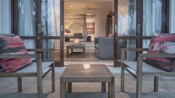 Ein modernes Wohnzimmer mit einem Holztisch, zwei Sesseln mit gemusterten Kissen und Blick in ein Schlafzimmer mit Himmelbett.