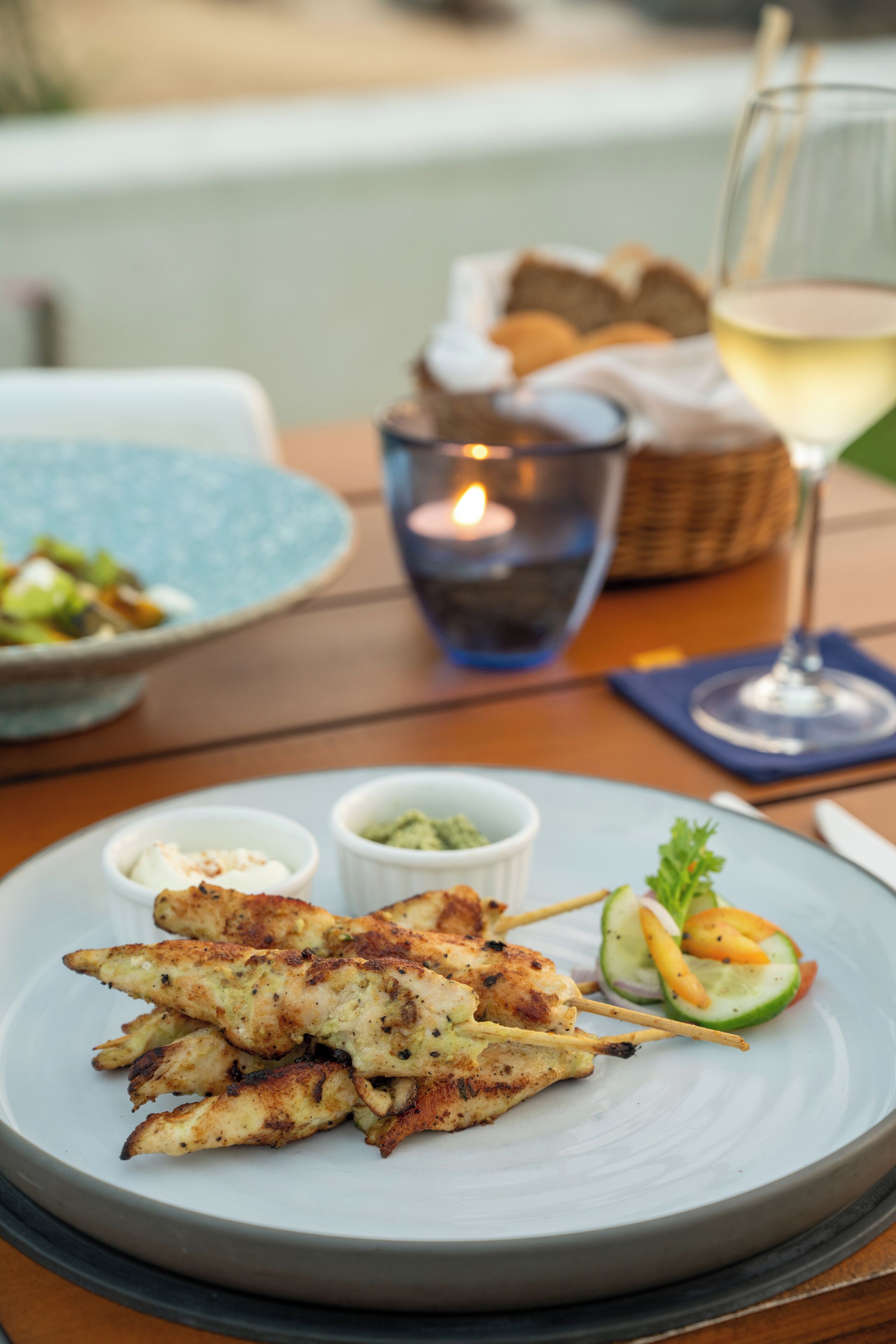 A plate of grilled chicken skewers with a side salad and a glass of white wine on a wooden table.