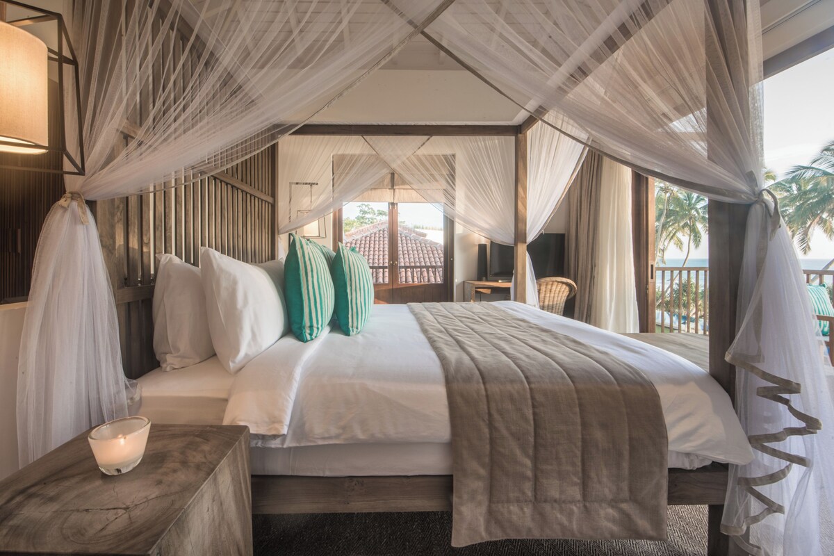 A bedroom with a canopy bed, a bedside table with a candle, and a view of the outside.