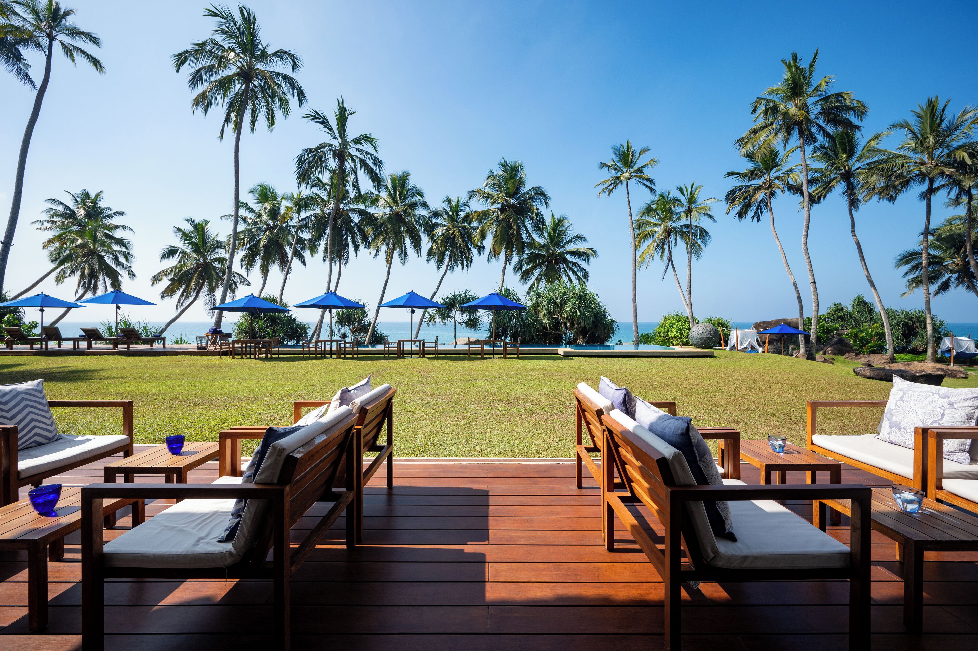 Terraza de madera con mobiliario exterior, palmeras y sombrillas con vistas a la playa.