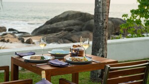 A wooden table set with plates of food, glasses of wine, and a candle, overlooking a rocky beach and ocean. - Kumu Beach (Balapitiya)