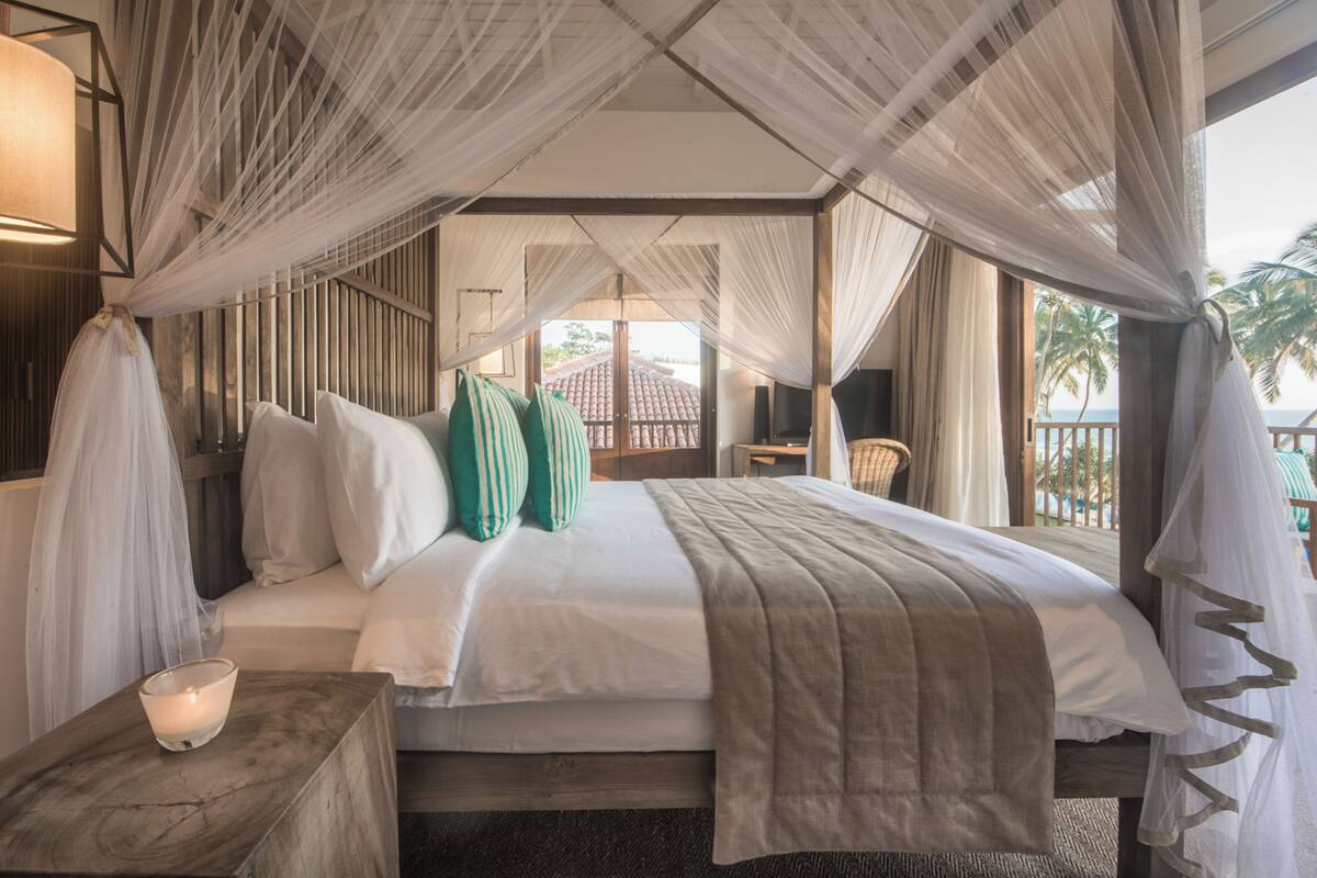A bedroom with a canopy bed, a bedside table with a candle, and a view of the outside.