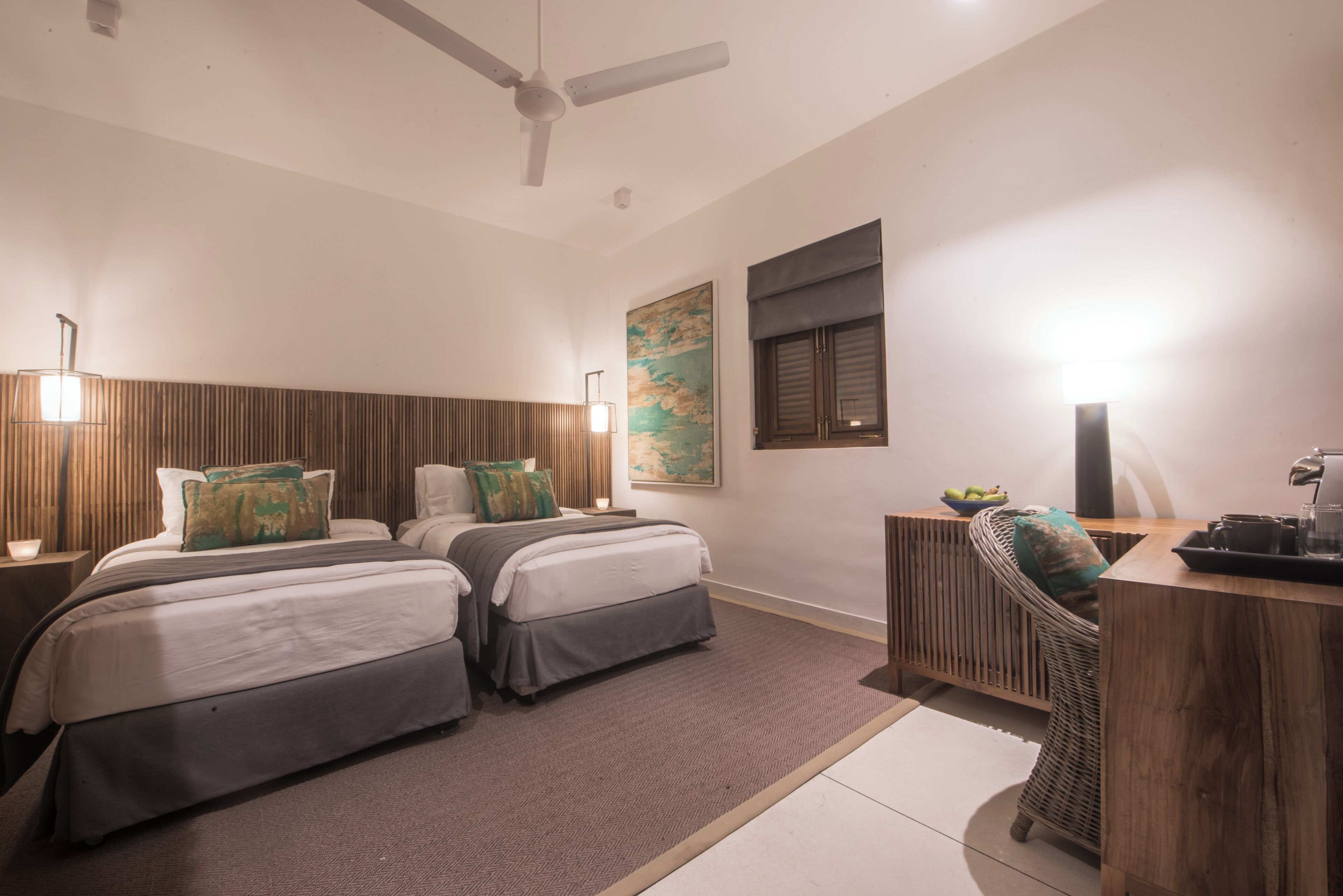 Habitación de hotel con dos camas, un escritorio, una silla, un ventilador de techo y una obra de arte en la pared.