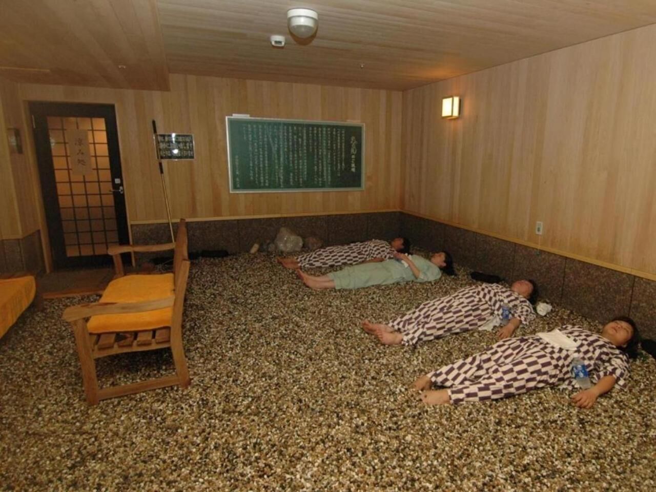 A sauna room with wooden walls and a floor covered in pebbles. There are three people lying on the floor, a wooden chair, and a sign on the wall.