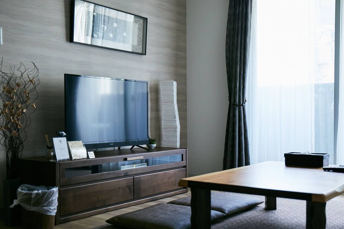 Una sala de estar moderna con una televisión de pantalla plana sobre un mueble de madera, una mesa de comedor con bancos y un ventanal con cortinas.