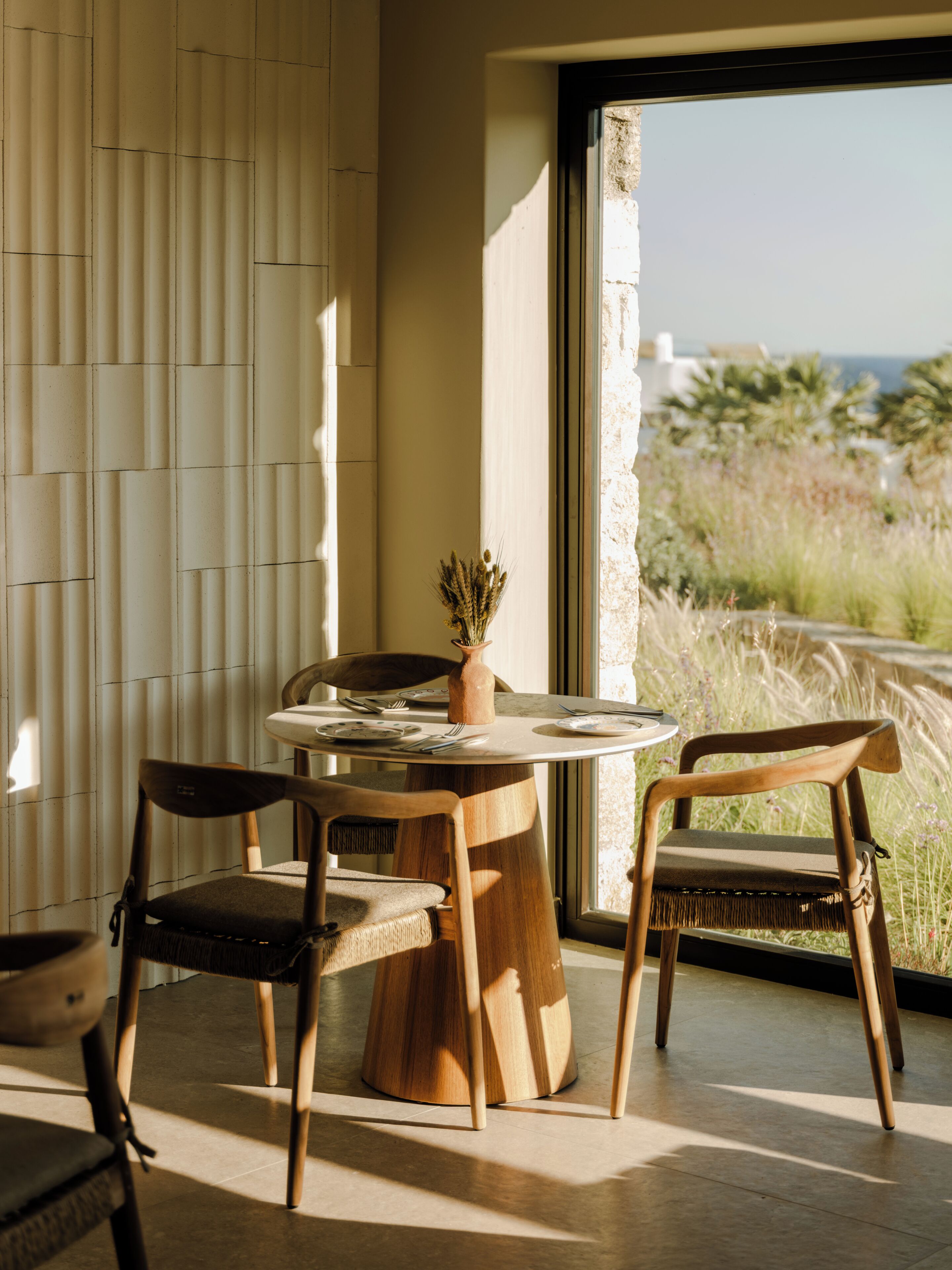 Un comedor con una mesa redonda, dos sillas, un florero con plantas secas y vistas al exterior a través de un ventanal.