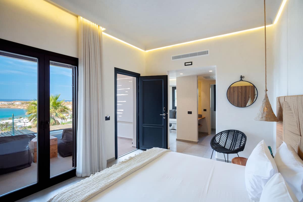 A modern hotel room with a large bed, a balcony with a view of the sea, and a separate sitting area with a chair and a table.