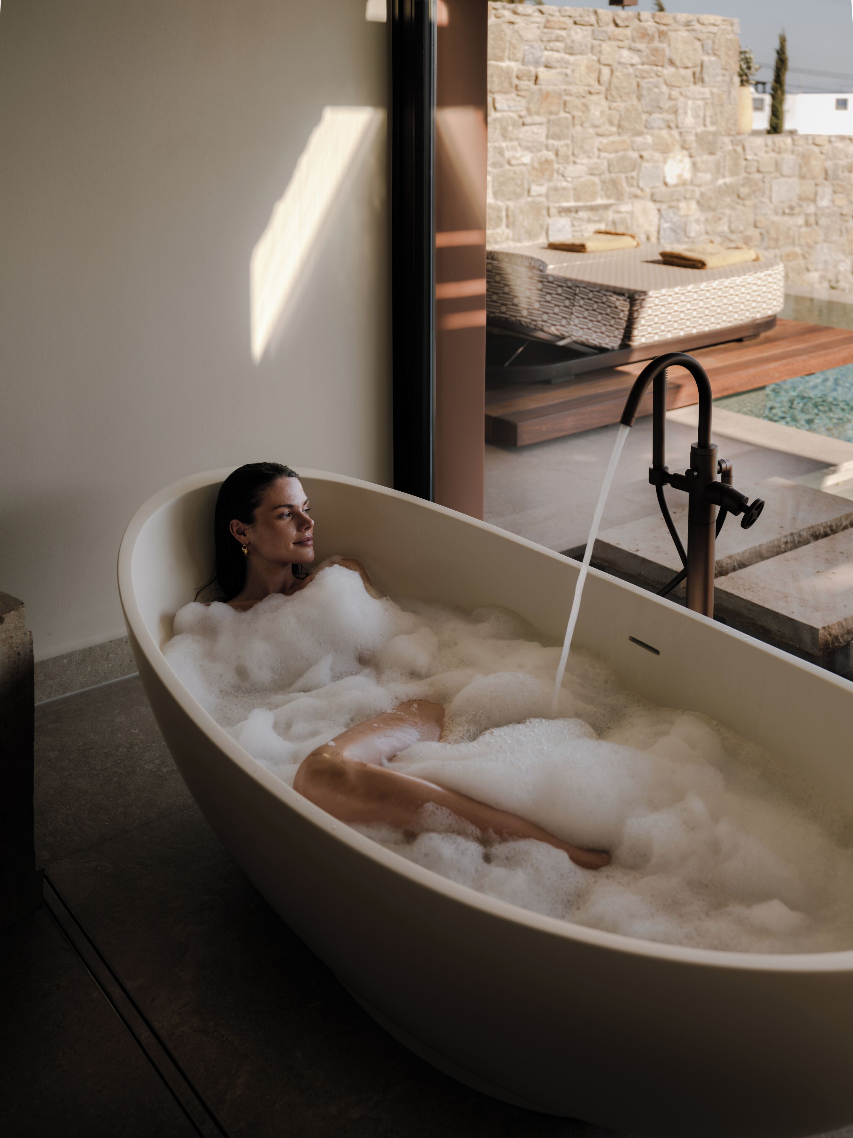 Une personne se détend dans une grande baignoire remplie de mousse, avec un mur de pierre et un espace de détente extérieur visibles en arrière-plan.