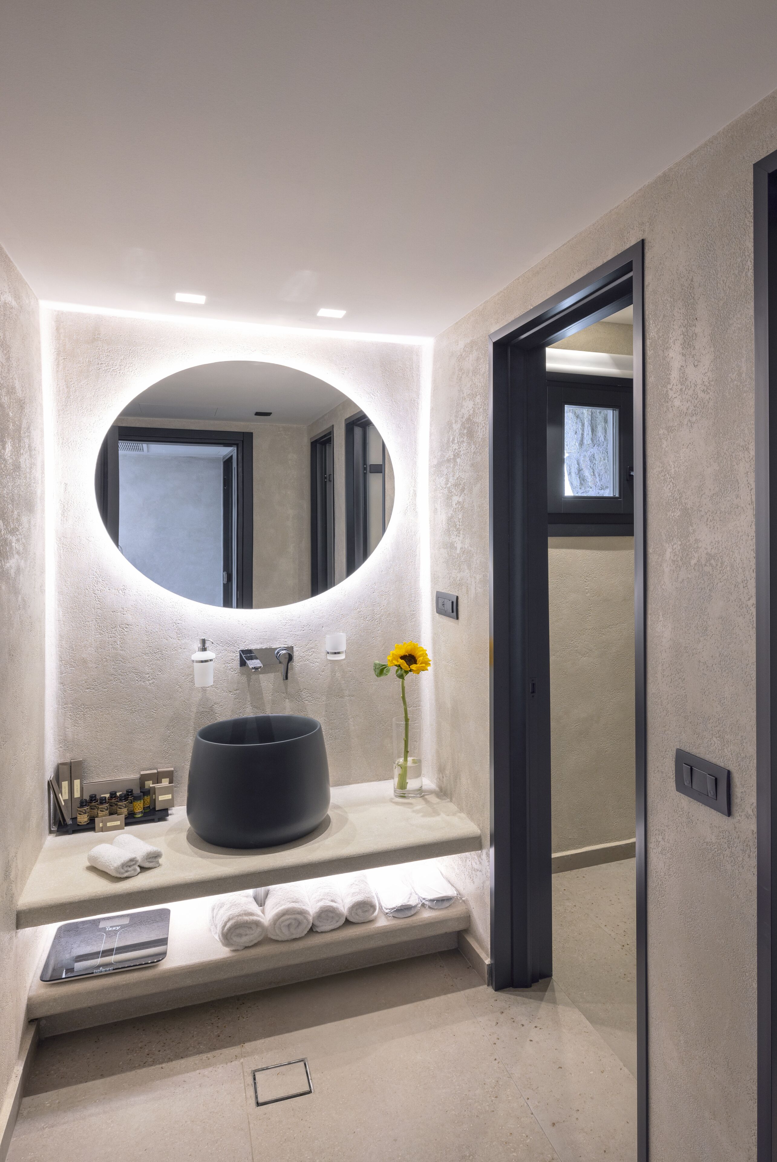 Une salle de bain moderne avec un miroir rond, un lavabo noir et un vase contenant une fleur jaune.