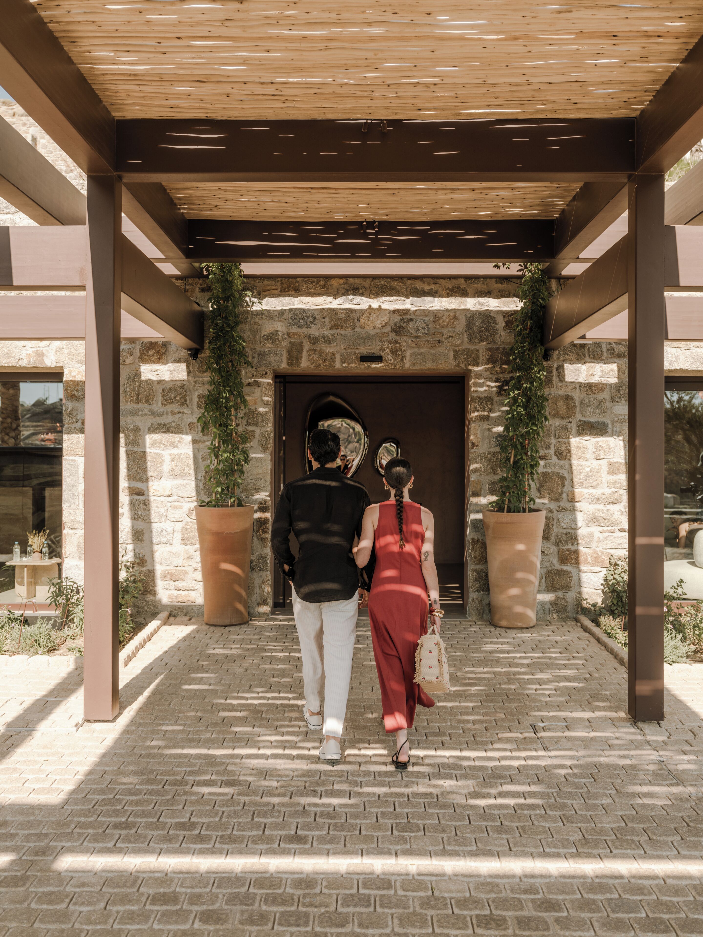 A couple walking towards a building entrance with a stone facade and large potted plants.
