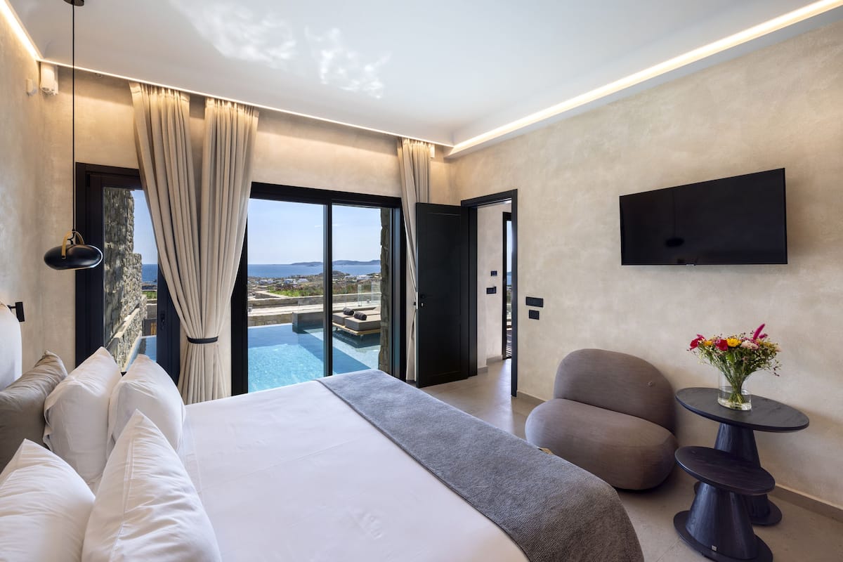 A modern hotel room with a large bed, a seating area, a TV, and a view of the sea.