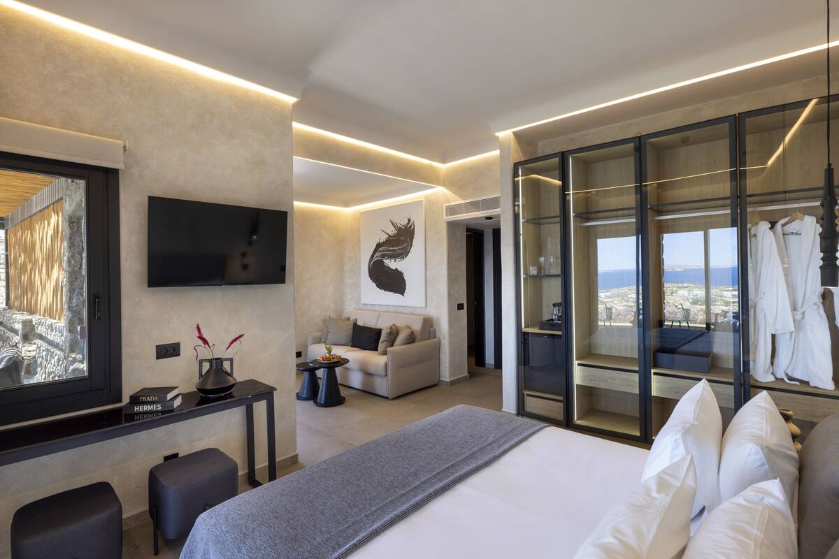 A modern hotel room with a large bed, a desk with a flat-screen TV, a sofa, and a glass-enclosed walk-in closet with robes.