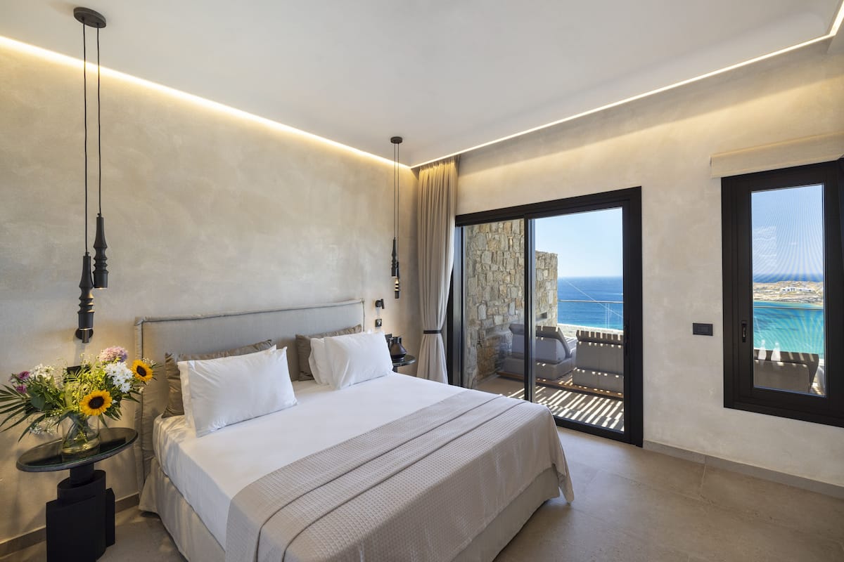 A modern hotel room with a large bed, a nightstand with a vase of flowers, a view of the ocean through a sliding glass door, and a balcony with a pool.