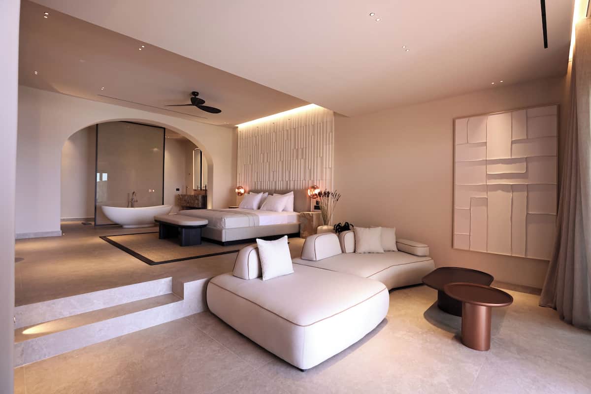 A modern, well-lit living area with a large sofa, a coffee table, and a seating area with a small table. There is a large mirror and a ceiling fan.