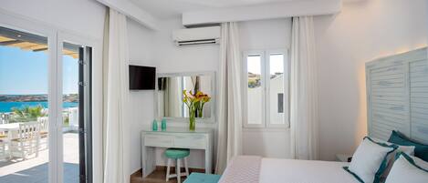 A bedroom with a bed, a desk, a chair, a TV, and a view of the sea.