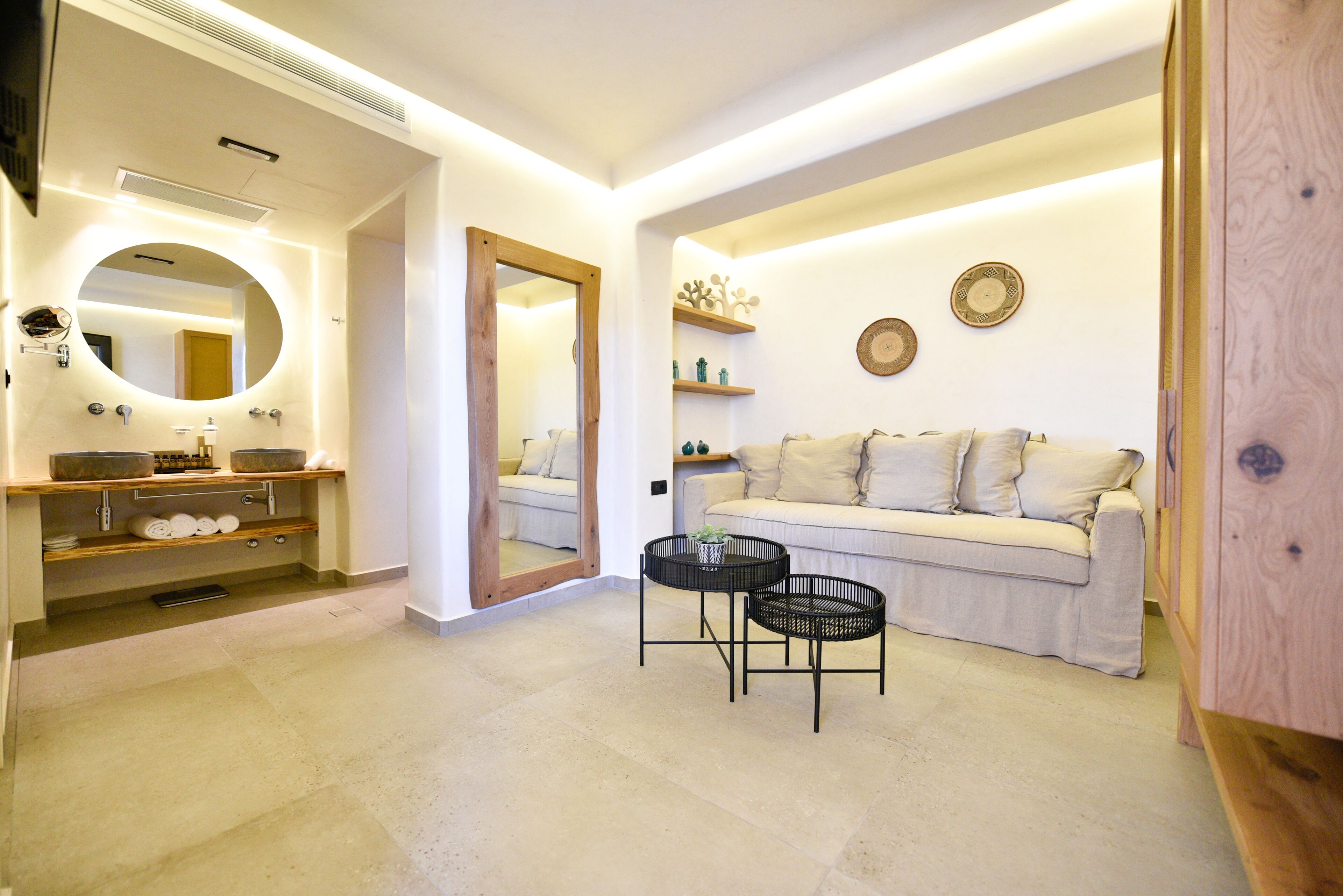 A modern bathroom with a vanity, mirror, and shelves. There is a sofa and a small table in the adjacent living area.