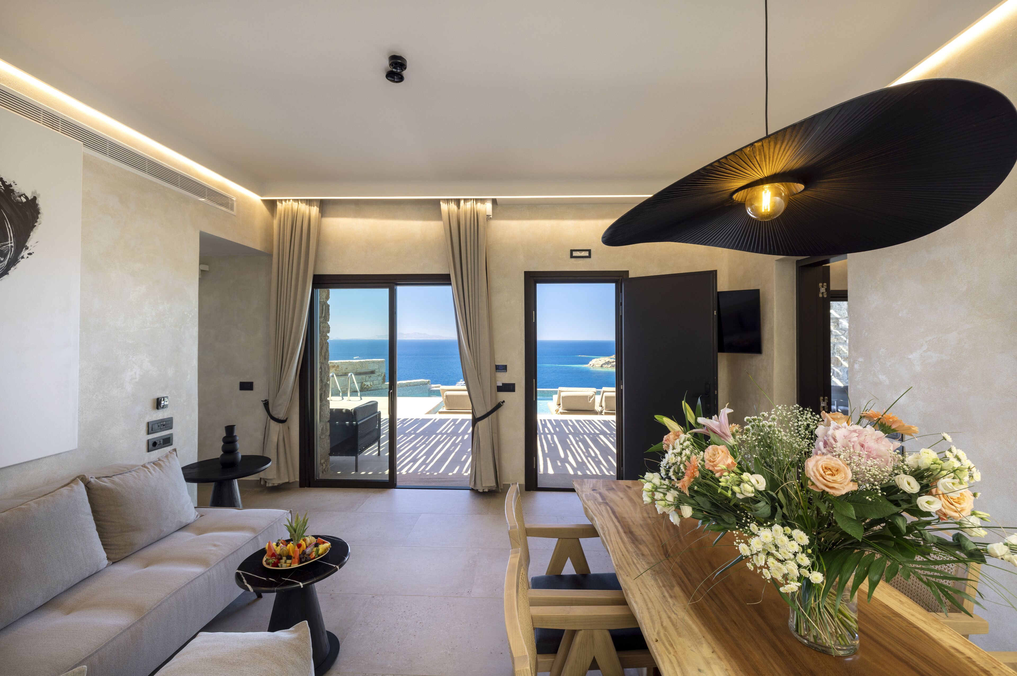 A modern living room with a sofa, a dining table with a vase of flowers, a view of the sea through large windows, and a ceiling light fixture.