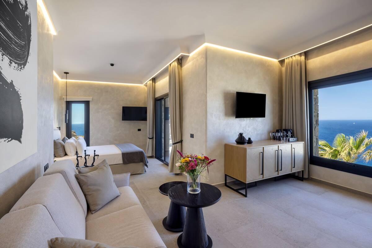 A modern hotel room with a sofa, a dining table with a vase of flowers, a minibar, a TV, and a large window with a view of the sea.